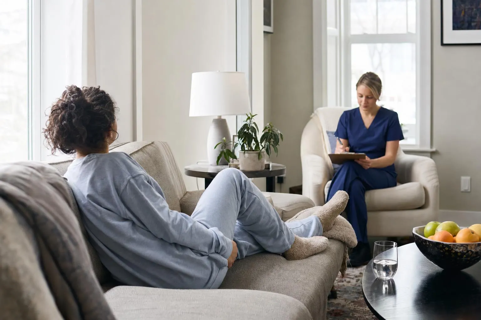 Patient in loungewear resting on a sofa at home with water and a light meal on a side table as a nurse checks in